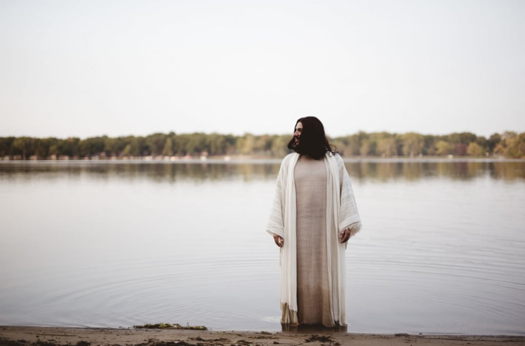 man with long hair standing in the water near the shore while looking in the distance